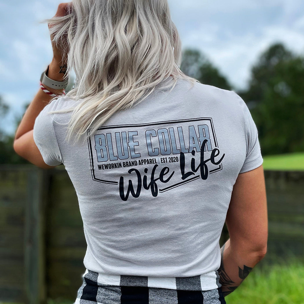 Woman outdoors, pictured from back, wearing a WE WORKIN Women’s short sleeve fitted tee in Silver color. Tee back is printed with the WW Blue Collar Wife Life graphic in black/grey ink. 