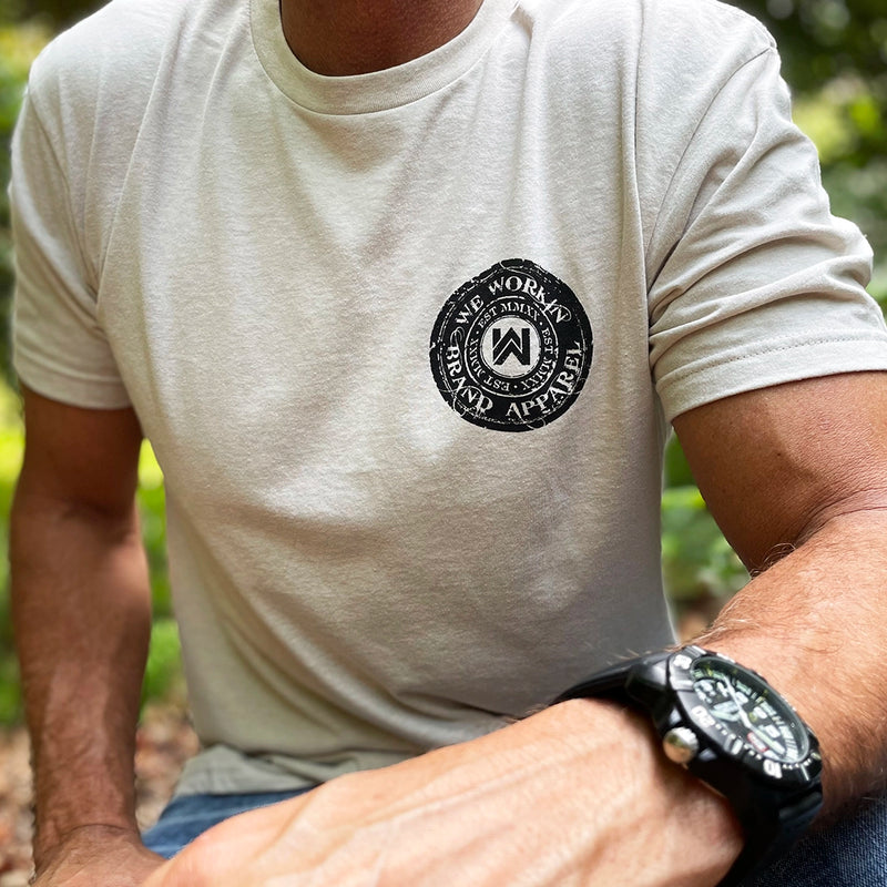 Man wearing a WW Sand color Graphic Tee (pictured from front). Circular graphic "WE WORKIN BRAND APPAREL" circular text and WW icon logo and EST MMXX smaller (also circular) inside the main text design in center of circular image. Printed small on the left chest "pocket" area in black ink.