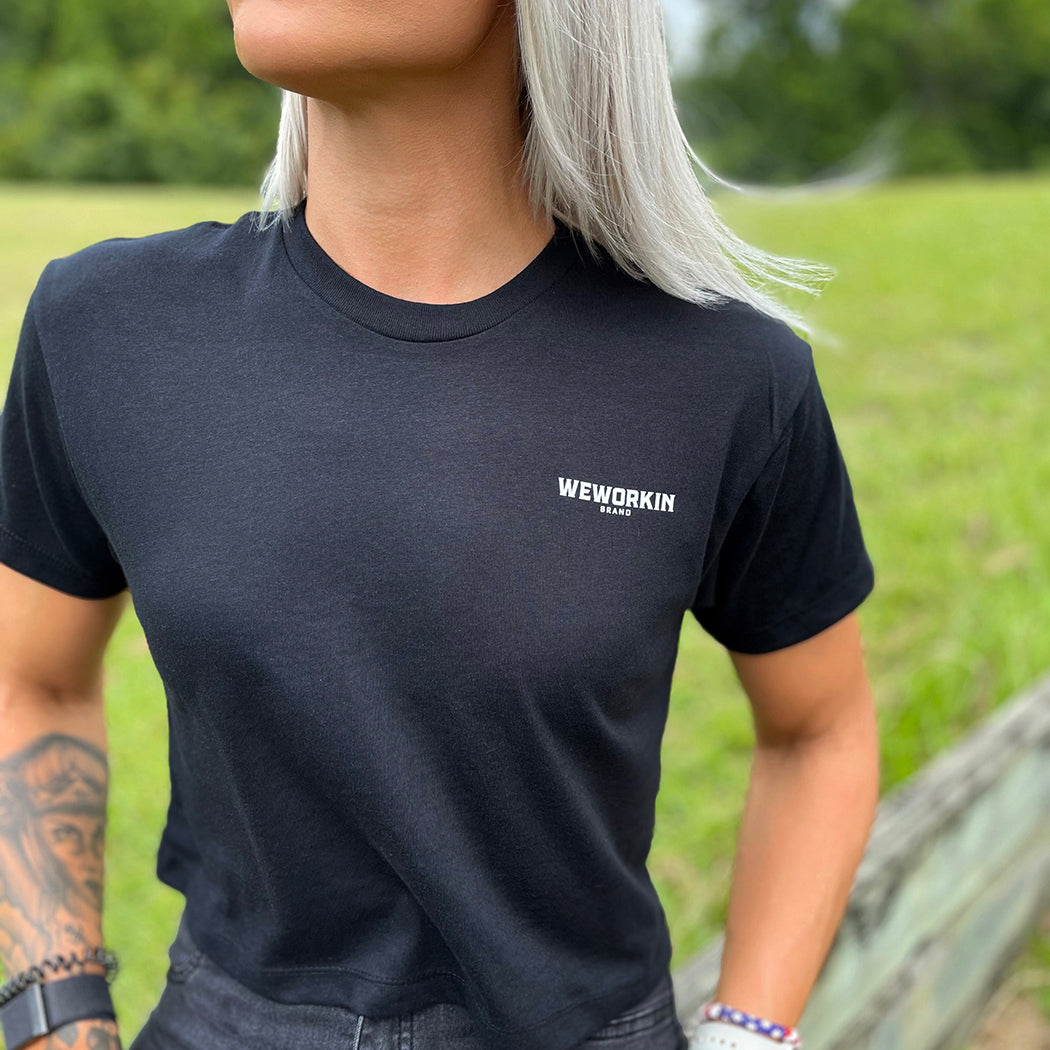 Front of a woman wearing a We Workin Women's short-sleeve cropped tee in black—with a small imprint of our "WEWORKIN BRAND" icon on the upper left chest in White ink.