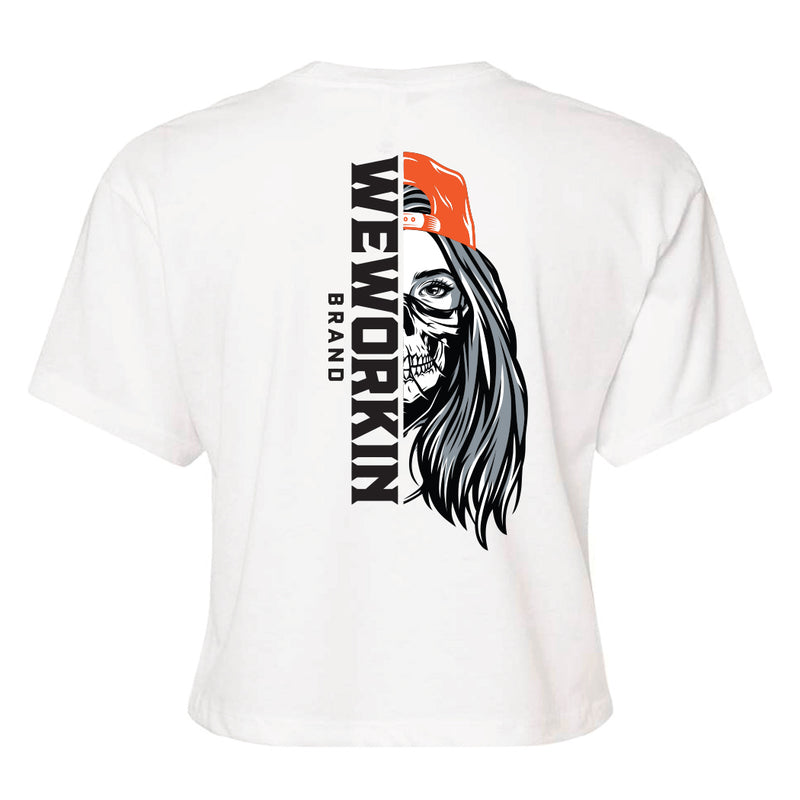Back of a We Workin Women's short-sleeve cropped tee in white—with a large imprint of our "WEWORKIN BRAND vertical text and Half Skull Woman with Hat" design in Black/White/Grey (hat graphic highlighted in Bright Orange ink.)