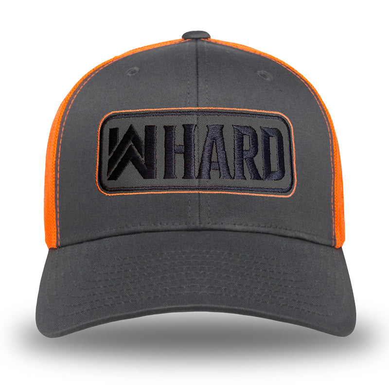 Front view of a Charcoal and Orange Retro Trucker hat. Embroidered with WW HARD "patch" style across the front (WW HARD icon/text in black thread, outer outline in orange thread, inner outline in black thread). Front 2 panels and bill are charcoal (matching charcoal undervisor), side/back mesh panels are orange. Pictured on white background