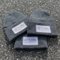 We Workin Dark Heathered Grey cuffed beanies laying on asphalt background. Custom 3.5" x 2" patches are sewn on the hats, with the WEWORKIN BRAND text and horizontal half-skull for the design on the patches—woven white background with grey thread for the design and white merrowed-edge border.