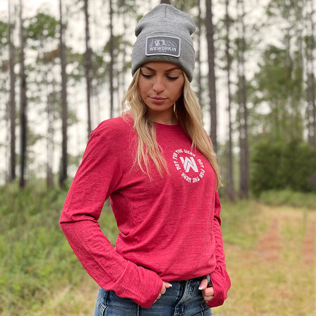 Woman pictured from front in heather red long-sleeved tee, imprinted with the We Workin icon centered on the chest with the text "Not For The Weak" encircling the icon in white ink. Also wearing a We Workin heather grey patch beanie.