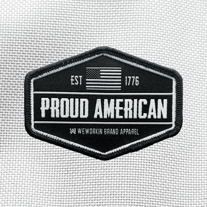 "PROUD AMERICAN" text centered large on velcro-back patch (both the hook and loop velcro sides included). [2] thread colors for the design (grey and white) on a black woven background, with black merrowed border. EST 1776 and US flag centered at top of patch.3.5" wide Woven patch displayed on a grey canvas background.