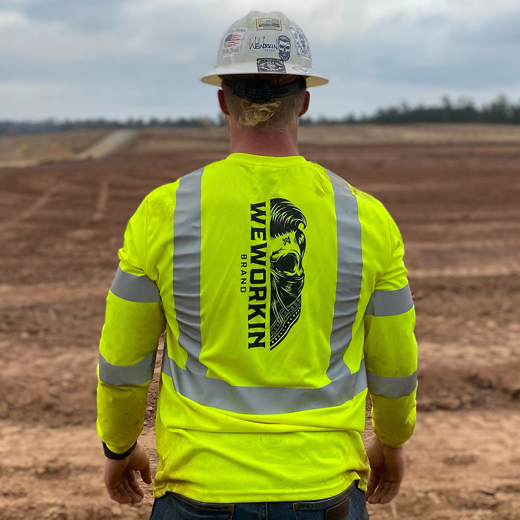 WORKWEAR Collection. Man pictured from back on jobsite wearing a hi-viz WW CLASS 3—Mesh Long Sleeve Tee with WWB Half Skull design and hard hat with WW hat stickers.