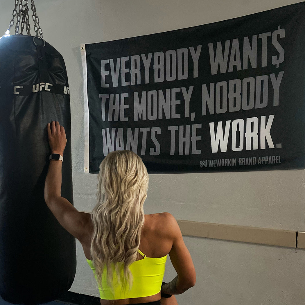 EXTRAS collection. 5' x 3' Flag with the We Workin quote "Everybody Wants the Money, Nobody Wants the WORK" in large grey/white letters on black background. 2 brass colored grommets. Hanging on the wall of a gym with a woman working out on a heavy bag.