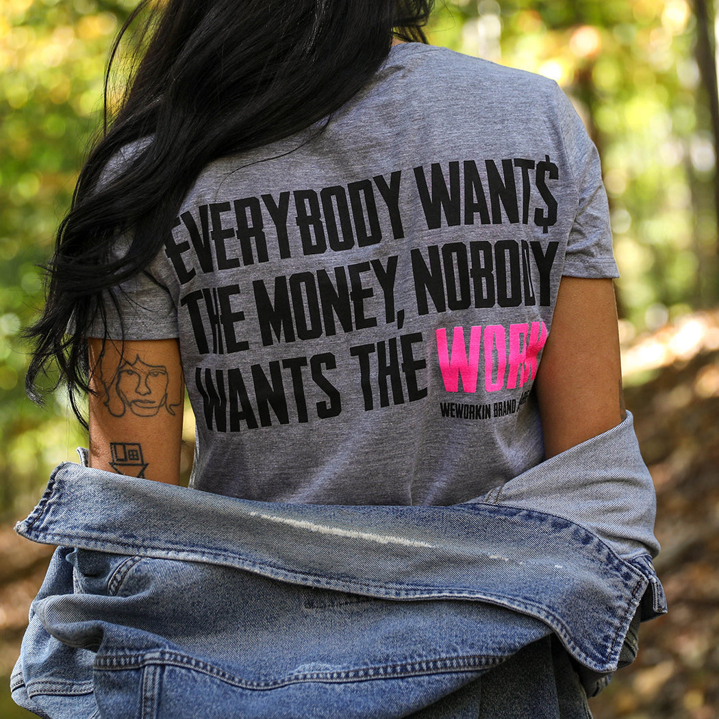 WOMEN's Collection. Woman is wearing a Grey tee with the tagline "Everybody Want$ the Money. Nobody Wants the WORK." printed on the back in white/pink inks.