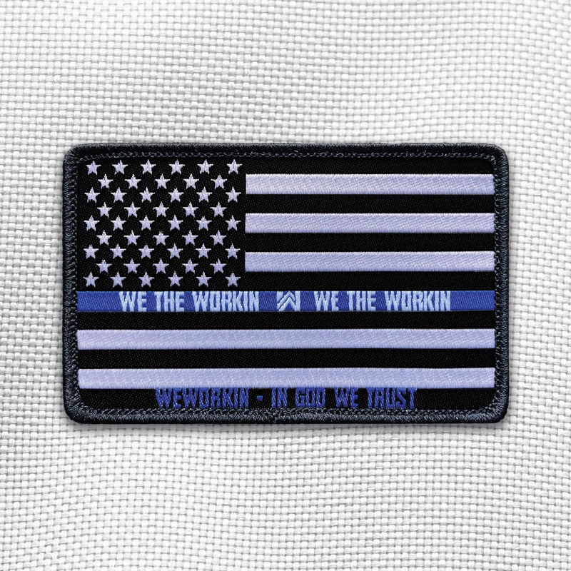 THIN BLUE LINE Flag on a velcro-backed patch (both the hook and loop sides provided). [1] thread color for the blue line stripe and the WEWORKIN • IN GOD WE TRUST  (white thread for remaining stripes, stars and WE THE WORKIN WW text) on a black woven background, with black merrowed border. 3.5" wide Woven patch displayed on a light grey canvas background.