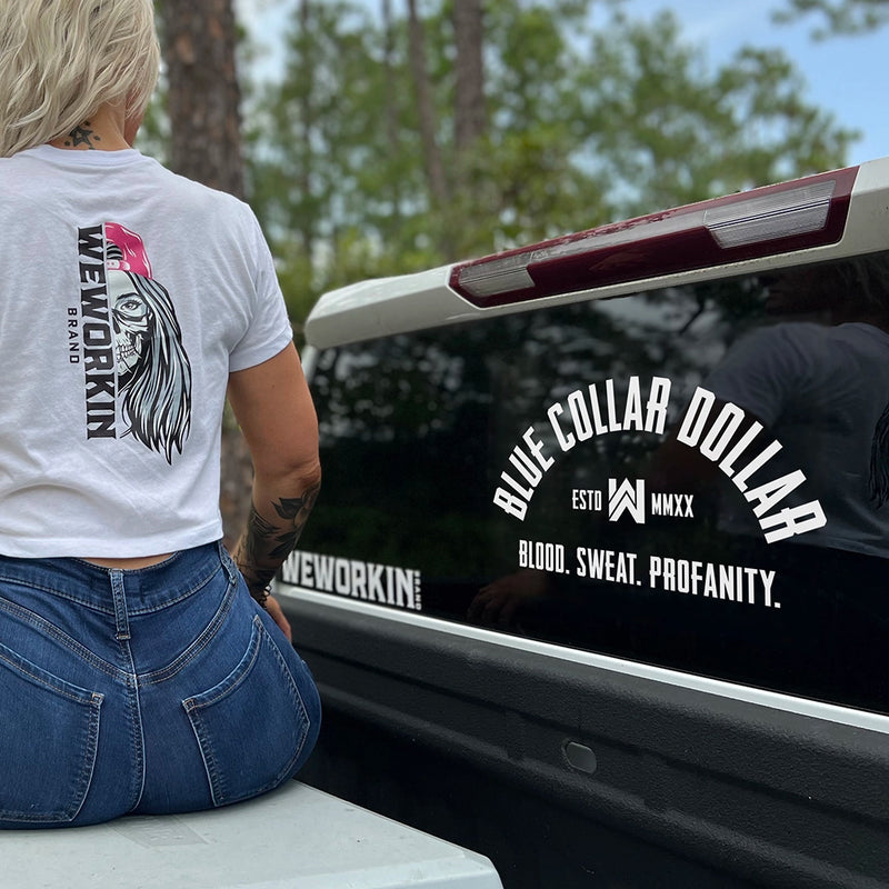 Large WEWORKIN BRAND "BLUE COLLAR DOLLAR" curve-design, white Decal—Custom die-cut Direct Transfer decal/sticker on the rear window of a pickup truck. Woman wearing a crop white WW-half-skull tee.