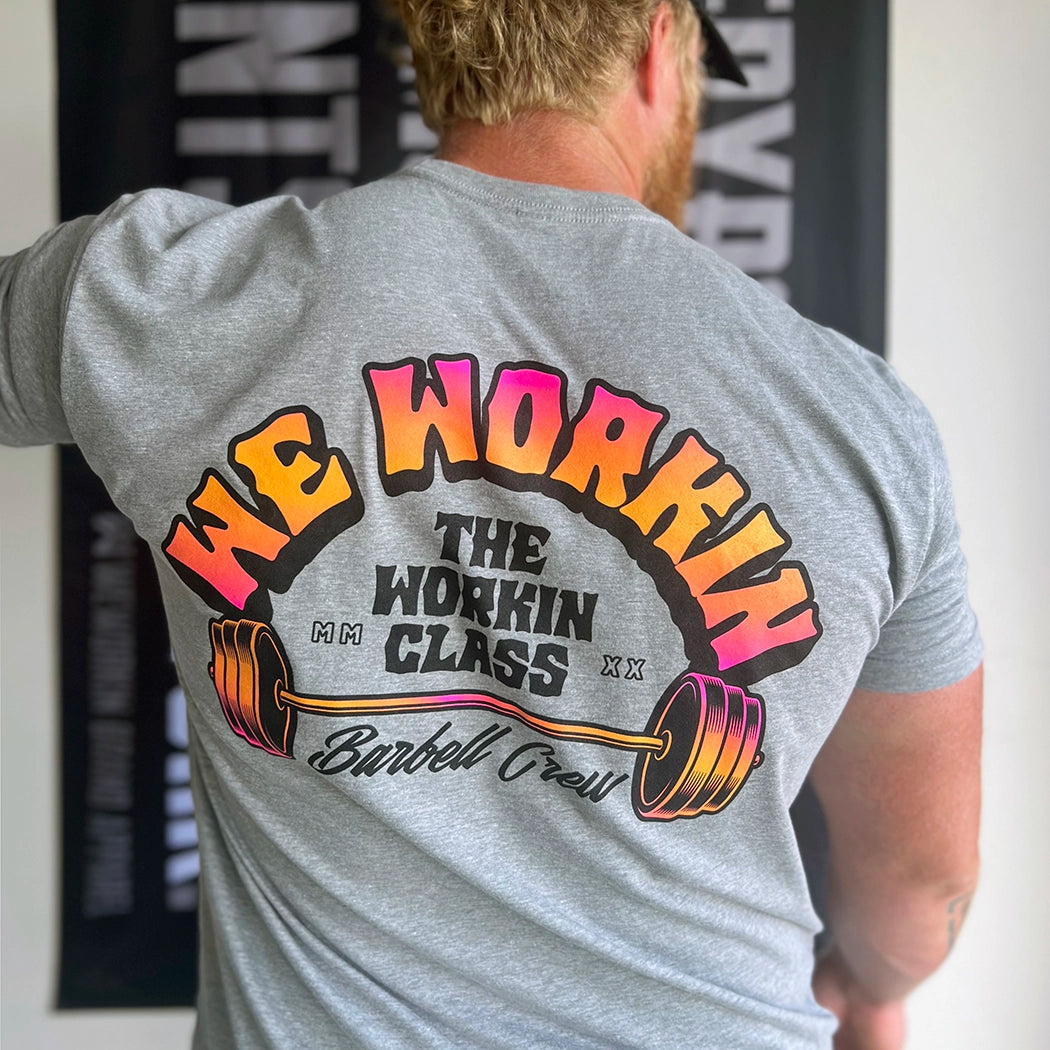 Man from back, wearing a WW heather grey tee. 3-color BARBELL CREW design in Neon Pink to Bright yellow fade and black, "WE WORKIN" main text is arched over top of "THE WORKIN CLASS" text and "Barbell Crew" script across the bottom—a Barbell in the center—design is printed large in the center/upper back.