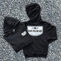 Two Blue Collar Kid black youth hoodies on a gravel surface (shows front and back). Imprint on the back is large and a small imprint on the left front "pocket" area. Design says "Blue Collar Kid. Just. Like. Dad."