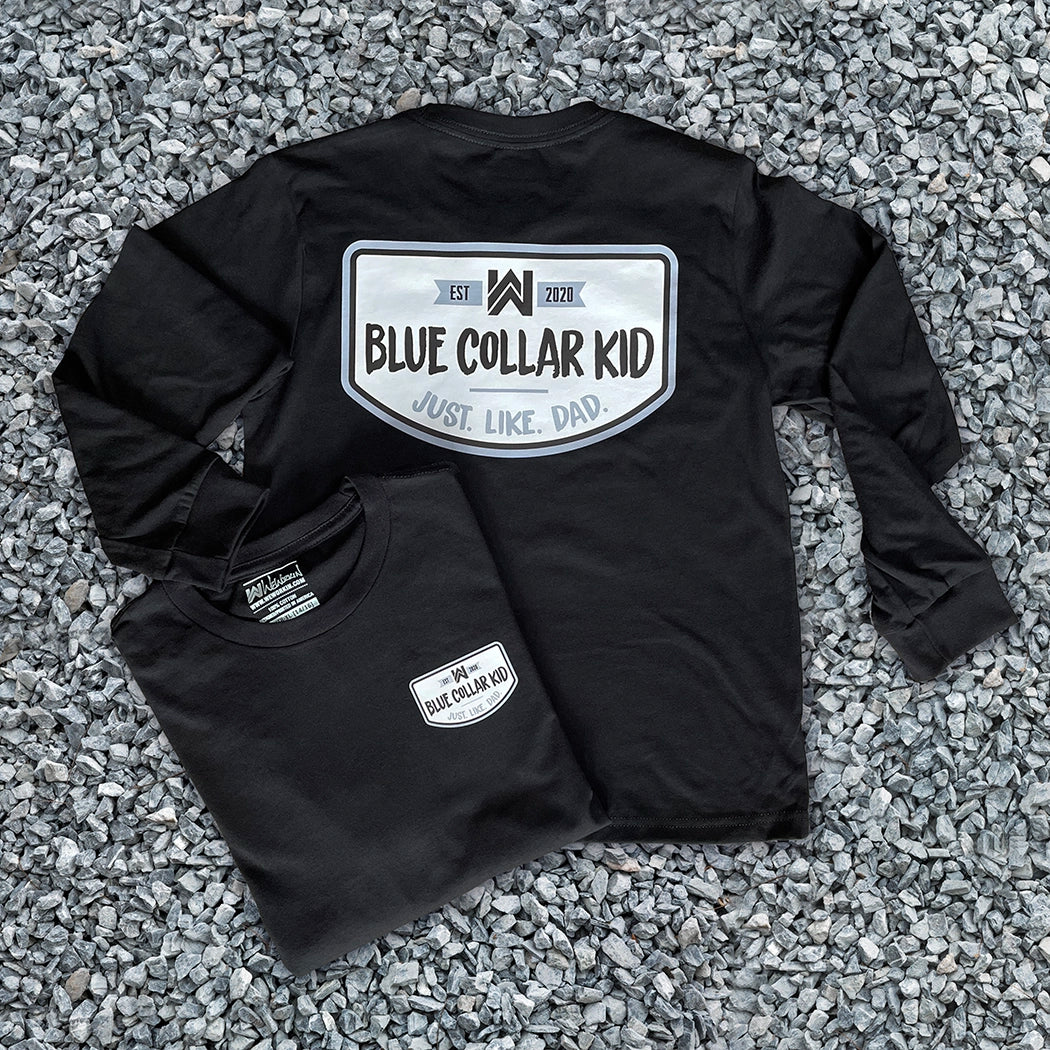 Two Blue Collar Kid black youth long sleeve tees on a gravel surface (shows front and back). Imprint on the back is large and a small imprint on the left front "pocket" area. Design says "Blue Collar Kid. Just. Like. Dad."