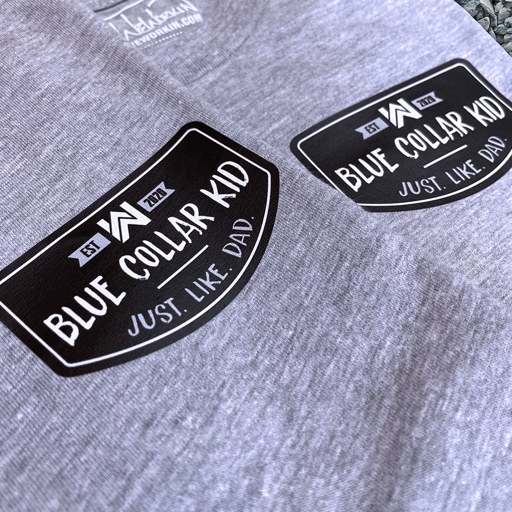 Two Blue Collar Kid grey youth long sleeve tees on a gravel surface (shows front). Small imprint on the left front "pocket" area. Design says "Blue Collar Kid. Just. Like. Dad."
