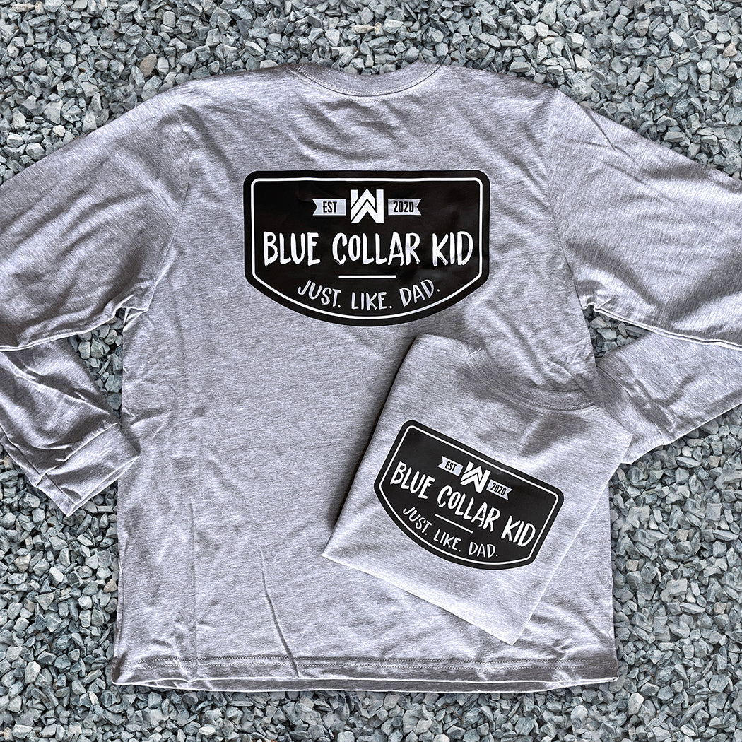 Two Blue Collar Kid grey youth long sleeve tees on a gravel surface (shows front and back). Imprint on the back is large and a small imprint on the left front "pocket" area. Design says "Blue Collar Kid. Just. Like. Dad."