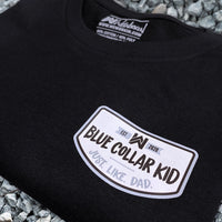 Blue Collar Kid black youth short sleeve tee on a gravel surface (shows front). Small imprint on the left front "pocket" area. Design says "Blue Collar Kid. Just. Like. Dad."