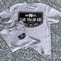 Two Blue Collar Kid grey youth short sleeve tees on a gravel surface (shows front and back). Small imprint on the left front "pocket" area. Design says "Blue Collar Kid. Just. Like. Dad."