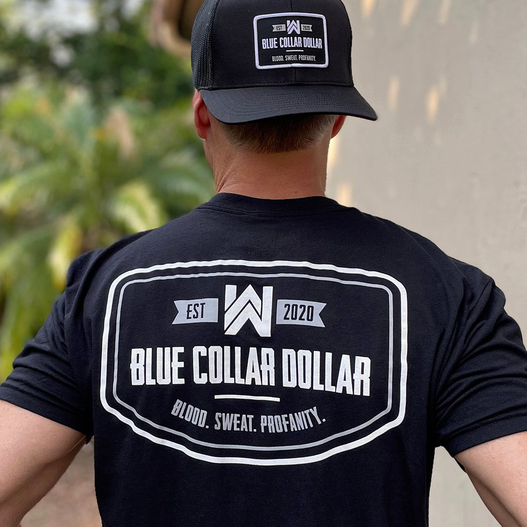 Male outdoors weaing a We Workin black, short sleeve tee with the BLUE COLLAR DOLLAR. BLOOD. SWEAT. PROFANITY. design, printed large on the upper back. Also wearing a Retro Classic 112 all black BCD patch hat.