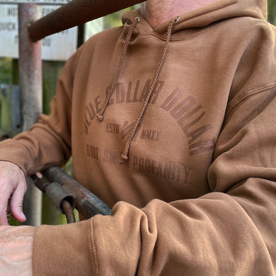 Man, outdoors, pictured close up wearing a WW "Blue Collar Dollar. Blood. Sweat. Profanity." graphic design on a brown hoodie, screenprinted in brown ink. "Stealth" effect.