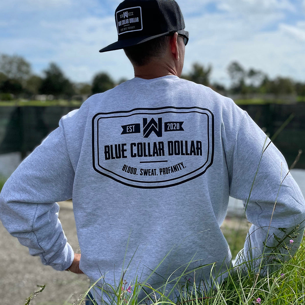Man outdoors pictured from back wearing a WW Ash Grey crewneck sweatshirt. "BLUE COLLAR DOLLAR—BLOOD. SWEAT. PROFANITY." printed across the full back in black ink. Banded cuffs and waist. (Also wearing a backwards WW Flatbill hat with large merrowed edge patch "BLUE COLLAR DOLLAR..."  in grey and white thread on woven black patch.)