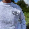 Close up of a man from the front, outdoors, wearing a WW Ash Grey crewneck sweatshirt. "BLUE COLLAR DOLLAR—BLOOD. SWEAT. PROFANITY." badge design printed on left chest "pocket" area in black ink.