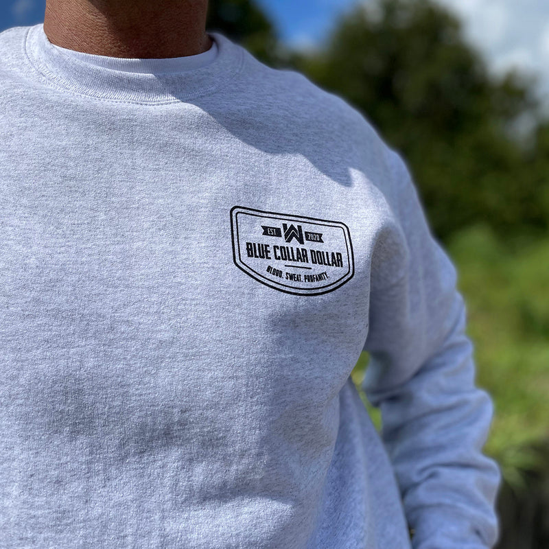 Close up of a man from the front, outdoors, wearing a WW Ash Grey crewneck sweatshirt. "BLUE COLLAR DOLLAR—BLOOD. SWEAT. PROFANITY." badge design printed on left chest "pocket" area in black ink.