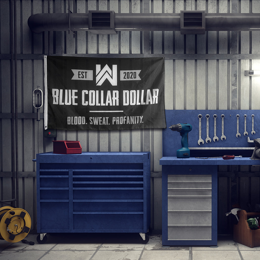 WeWorkin Brand "BLUE COLLAR DOLLAR—Blood. Sweat. Profanity." flag hung on a shop wall above a blue toolbox. Each flag measures approx. 5'w x 3'h, black background with grey/white letters. White, double-stitched, thicker left edge for durability, (2) grommets (one at top left and one at bottom left corners).