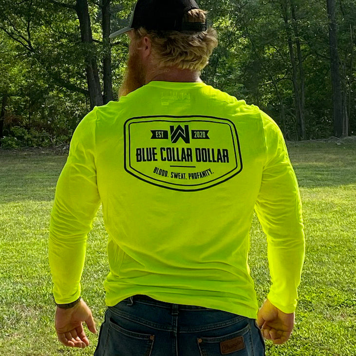 Man outdoors wearing a We Workin long sleeve hi-viz tee, with the BLUE COLLAR COLLAR. BLOOD. SWEAT. PROFANITY. design on the back. Also wearing a heather grey/black 112 classic trucker WW patch hat.