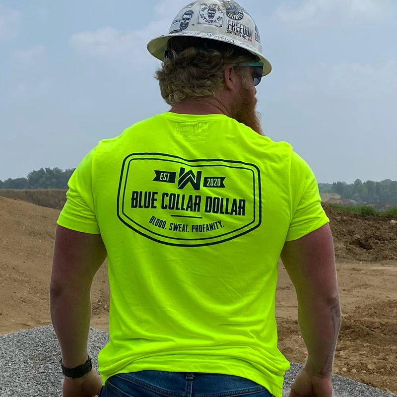 Man outdoors on construction site wearing a We Workin short sleeve hi-viz tee, with the BLUE COLLAR COLLAR. BLOOD. SWEAT. PROFANITY. design on the back. Also wearing a hard hat with WW stickers.