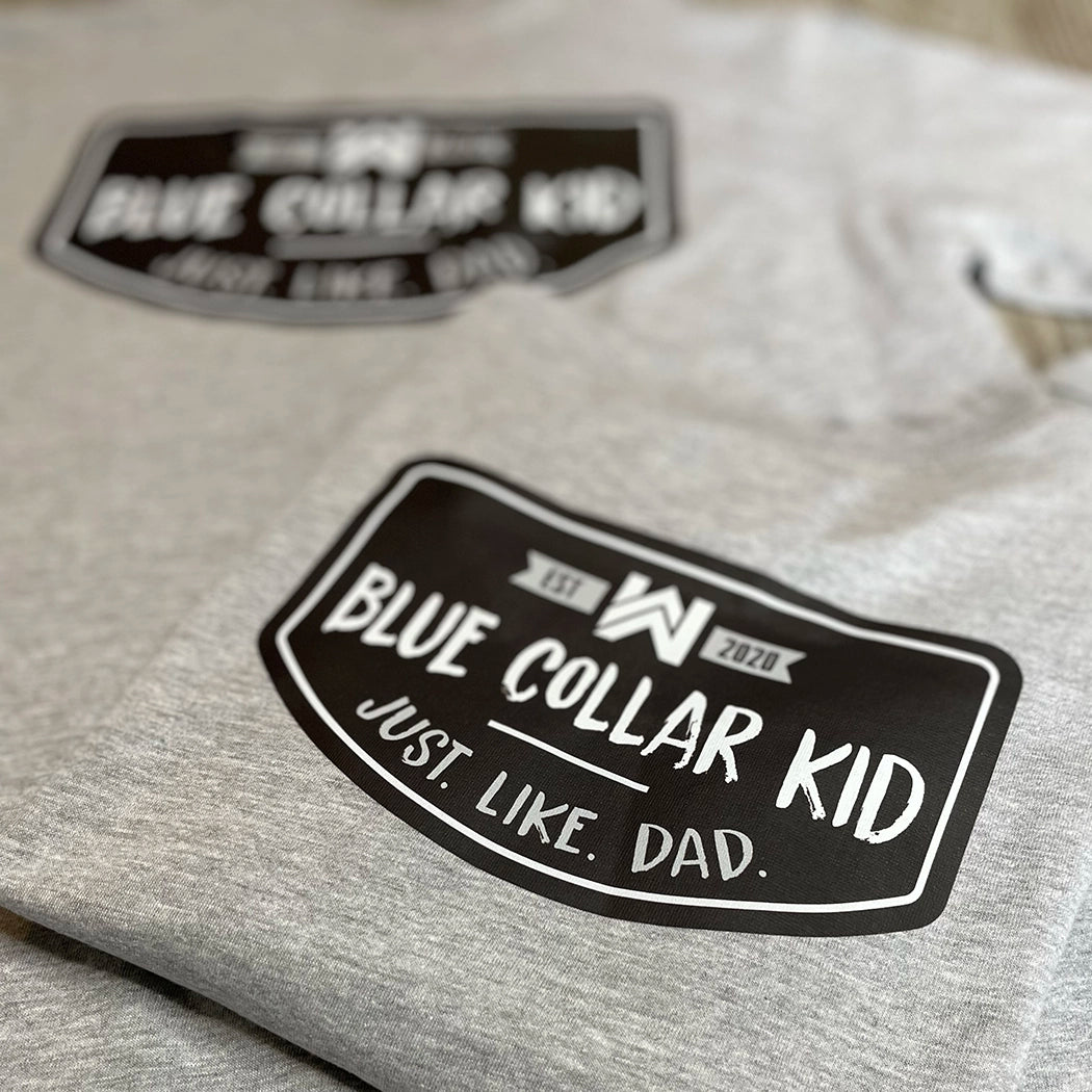 Two Blue Collar Kid grey youth short sleeve tees on a tile surface (shows back). Imprint on the back is large. Design says "Blue Collar Kid. Just. Like. Dad."