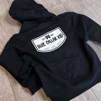 A Blue Collar Kid black youth hoodie on a tile background (shows back). Large design on back says "Blue Collar Kid. Just. Like. Mom." with WW icon and EST 2020.