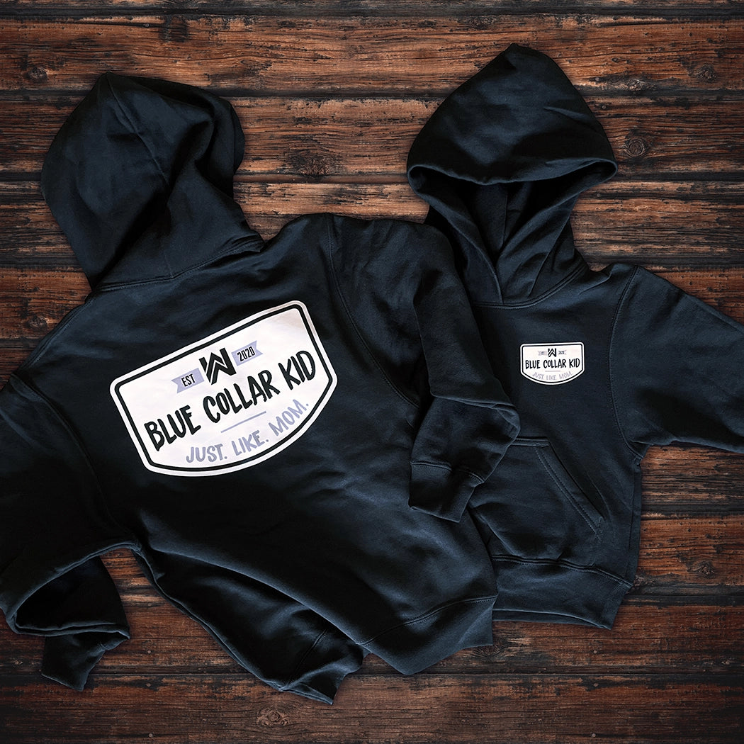 Two Blue Collar Kid black youth hoodies on a wood background (shows front and back). Large imprint on back, small imprint on the left front "pocket" area. Design says "Blue Collar Kid. Just. Like. Mom." with WW icon and EST 2020.