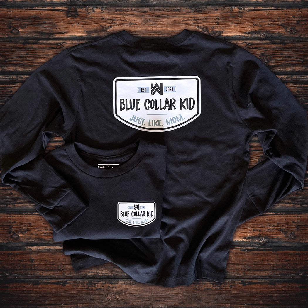 Two Blue Collar Kid black youth long sleeve tees on a wood background (shows front and back). Large imprint on back, small imprint on the left front "pocket" area. Design says "Blue Collar Kid. Just. Like. Mom." with WW icon and EST 2020.