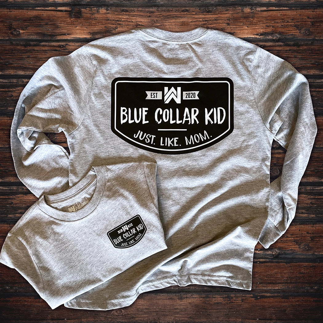 Two Blue Collar Kid heather grey youth long sleeve tees on a wood background (shows front and back). Large imprint on back, small imprint on the left front "pocket" area. Design says "Blue Collar Kid. Just. Like. Mom." with WW icon and EST 2020.