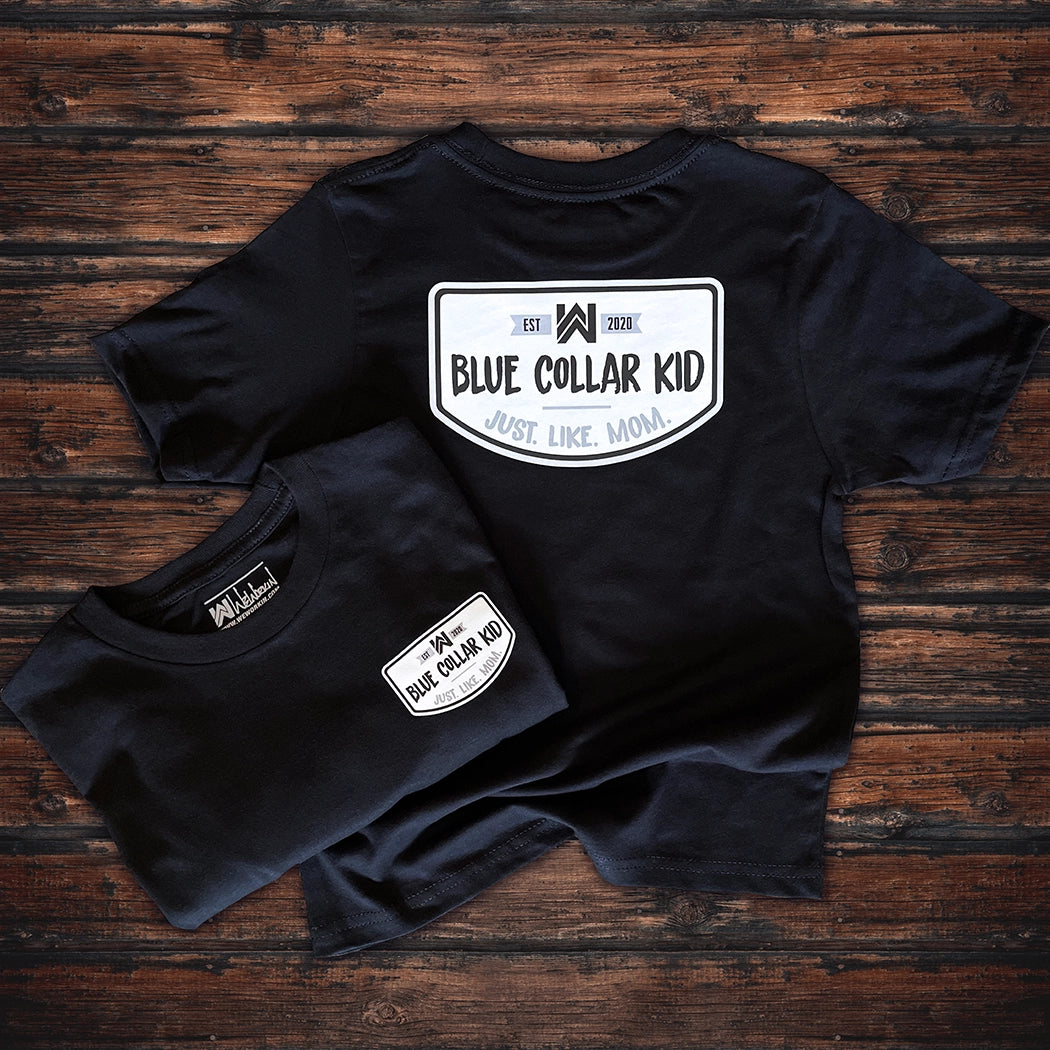 Two Blue Collar Kid black youth short sleeve tees on a wood background (shows front and back). Large imprint on back, small imprint on the left front "pocket" area. Design says "Blue Collar Kid. Just. Like. Mom." with WW icon and EST 2020.