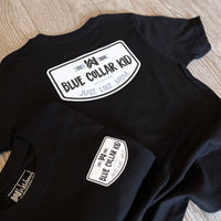 Two Blue Collar Kid black youth short sleeve tees on a tile background (shows front and back). Large imprint on back, small imprint on the left front "pocket" area. Design says "Blue Collar Kid. Just. Like. Mom." with WW icon and EST 2020.