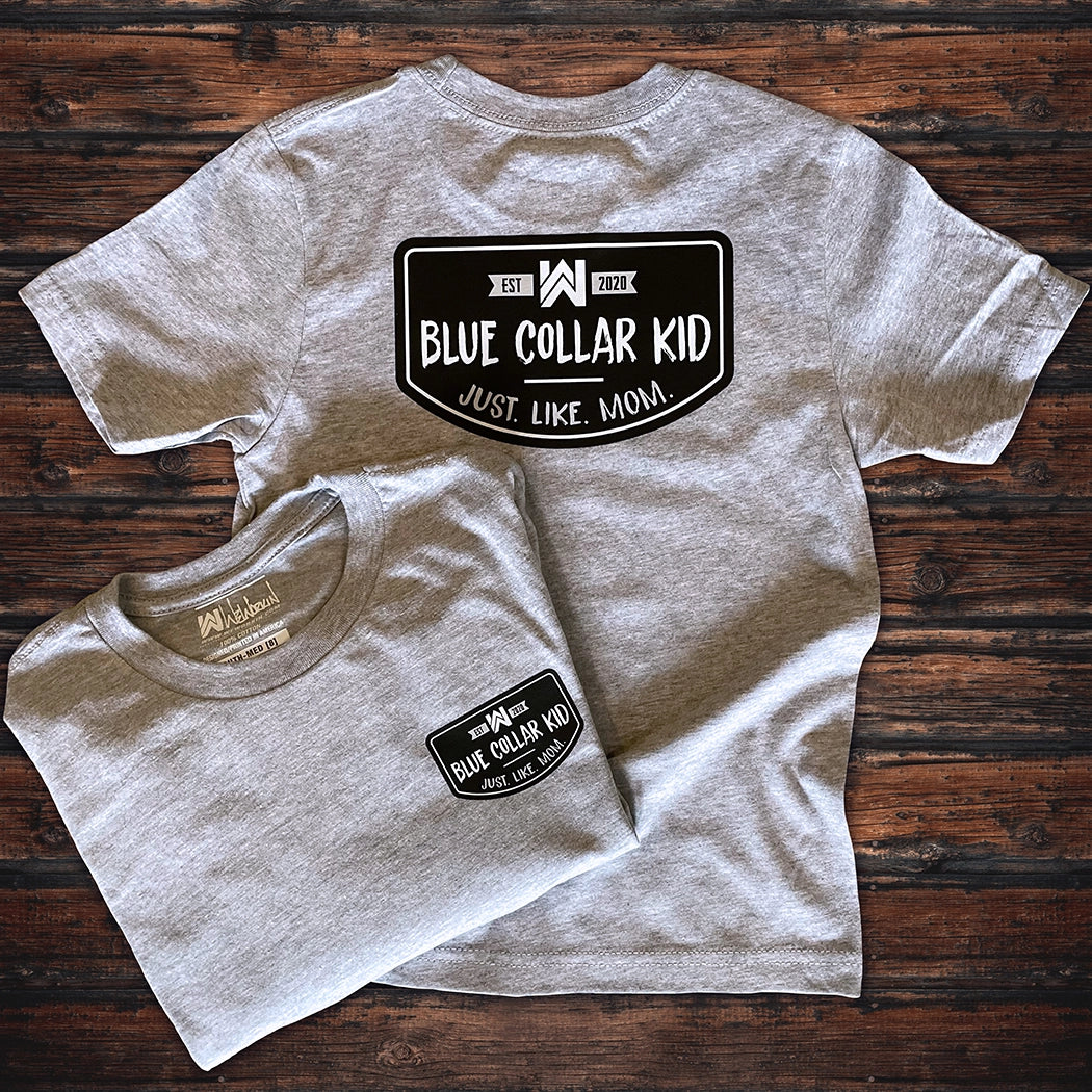 Two Blue Collar Kid heather grey youth short sleeve tees on a wood background (shows front and back). Large imprint on back, small imprint on the left front "pocket" area. Design says "Blue Collar Kid. Just. Like. Mom." with WW icon and EST 2020.