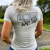 Woman outdoors, pictured from back, wearing jeans and a WE WORKIN Women’s short sleeve fitted tee in Silver color. Tee back is printed with the WW Blue Collar Wife Life graphic in black/grey ink. 