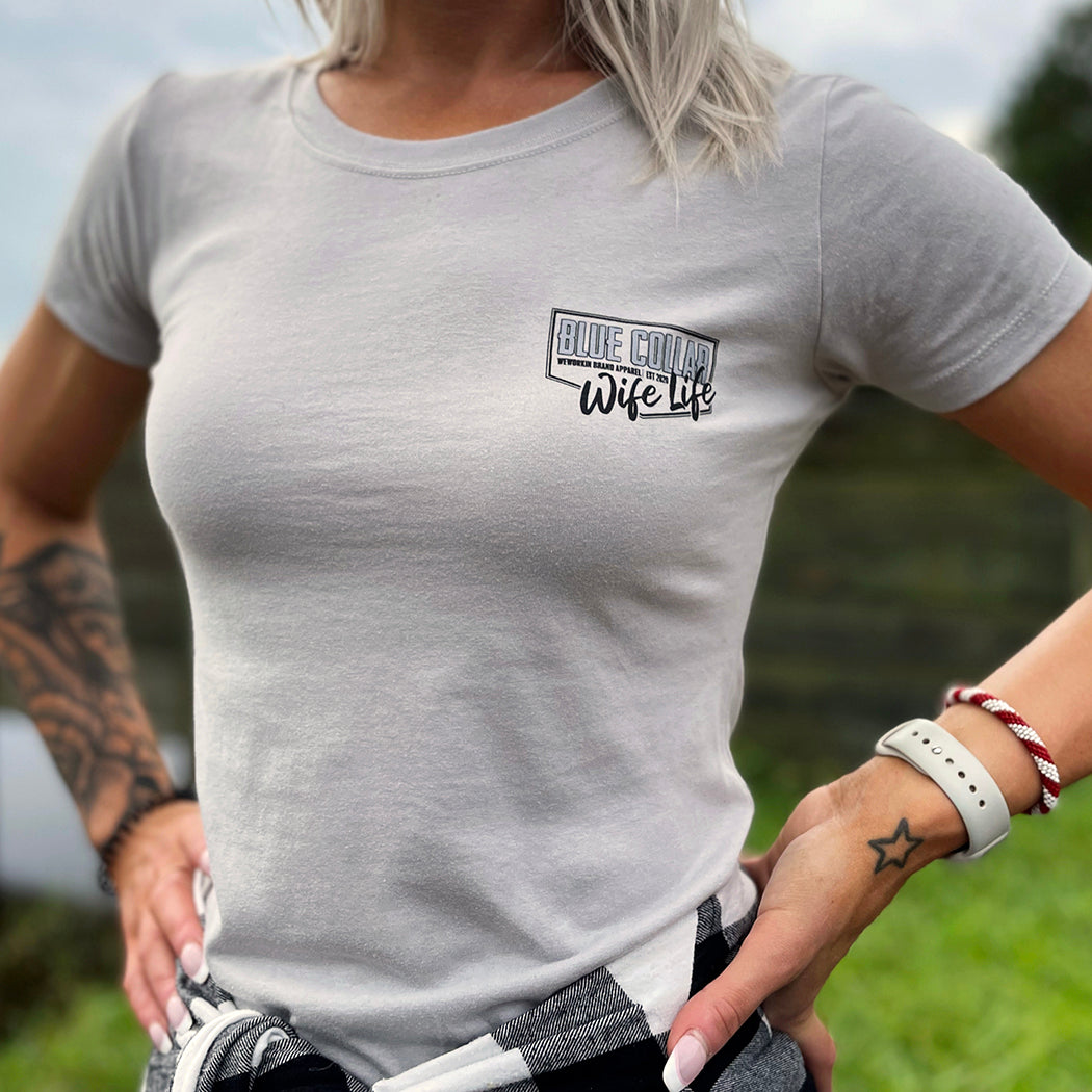 Woman outdoors, pictured from front, wearing a WE WORKIN Women’s short sleeve fitted tee in Silver color. Tee front "pocket" area is printed with the WW Blue Collar Wife Life graphic in black/grey ink. 