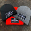 3 We Workin "BLUE COLLAR DOLLAR" cuffed patch beanies laying on a tree stump—available in 3 colors shown...Black, Heathered Grey and Neon Safety Orange. Custom woven BCD patches are sewn on the cuffs, merrowed-edge borders.