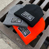 3 We Workin "EVERYBODY WANT$ THE MONEY..." cuffed patch beanies laying on a metal grate—available in 3 colors shown...Black, Heather Grey and Neon Safety Orange. Custom woven EWTM patches are sewn on the cuffs, merrowed-edge borders.
