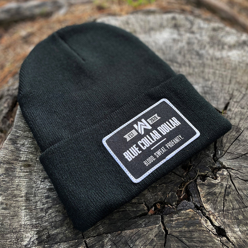 A We Workin Black "BLUE COLLAR DOLLAR" cuffed patch beanie laying on a tree stump. A custom woven BCD patch is sewn on the cuff, with merrowed-edge border.
