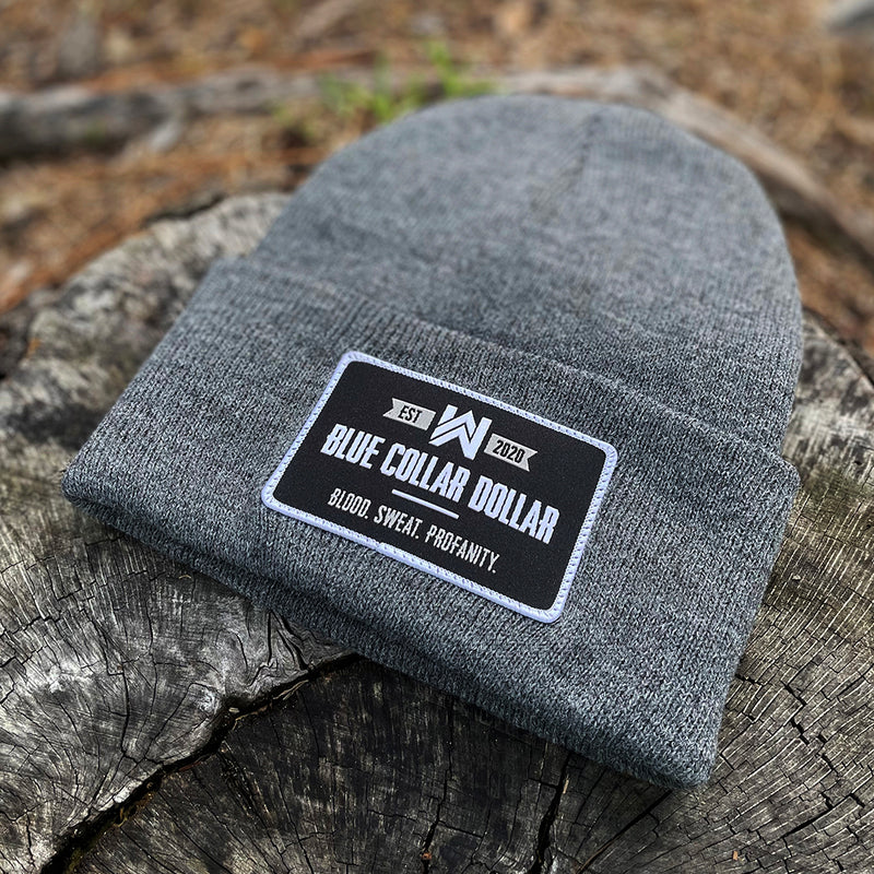 A We Workin Heathered Grey "BLUE COLLAR DOLLAR" cuffed patch beanie laying on a tree stump. A custom woven BCD patch is sewn on the cuff, with merrowed-edge border.