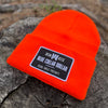 A We Workin Neon Safety Orange "BLUE COLLAR DOLLAR" cuffed patch beanie laying on a tree stump. A custom woven BCD patch is sewn on the cuff, with merrowed-edge border.