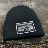 A We Workin Black "EVERYBODY WANT$ THE MONEY..." cuffed patch beanie laying on a tree stump. A custom woven EWTM patch is sewn on the cuff, with merrowed-edge border.
