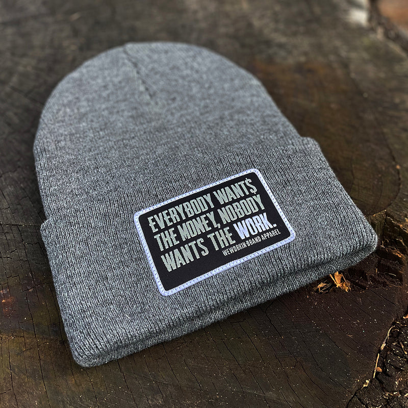 A We Workin Heathered Grey "EVERYBODY WANT$ THE MONEY..." cuffed patch beanie laying on a tree stump. A custom woven EWTM patch is sewn on the cuff, with merrowed-edge border.