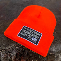 A We Workin Neon Safety Orange "EVERYBODY WANT$ THE MONEY..." cuffed patch beanie laying on a tree stump. A custom woven EWTM patch is sewn on the cuff, with merrowed-edge border.