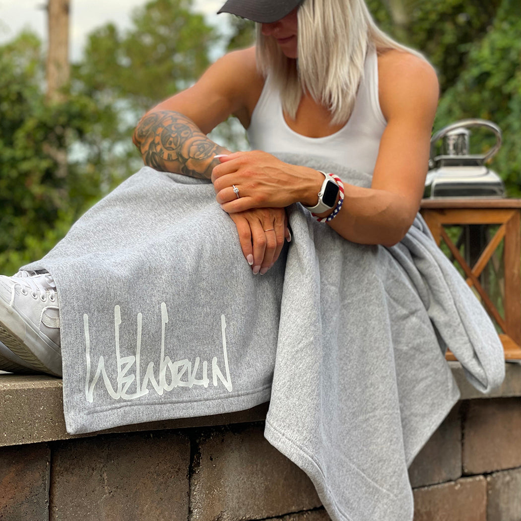 Woman sitting on stone wall with an oversized We Workin Athletic Grey Fleece Stadium blanket with Script logo-style draped over her.