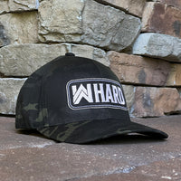 Front right view of Flexfit hat. MultiCam® black pattern fabric embroidered with WW HARD "patch" style across the front (WW HARD icon/text in white thread, outer outline in white thread, inner outline in grey thread). Front bill in MultiCam® black pattern.