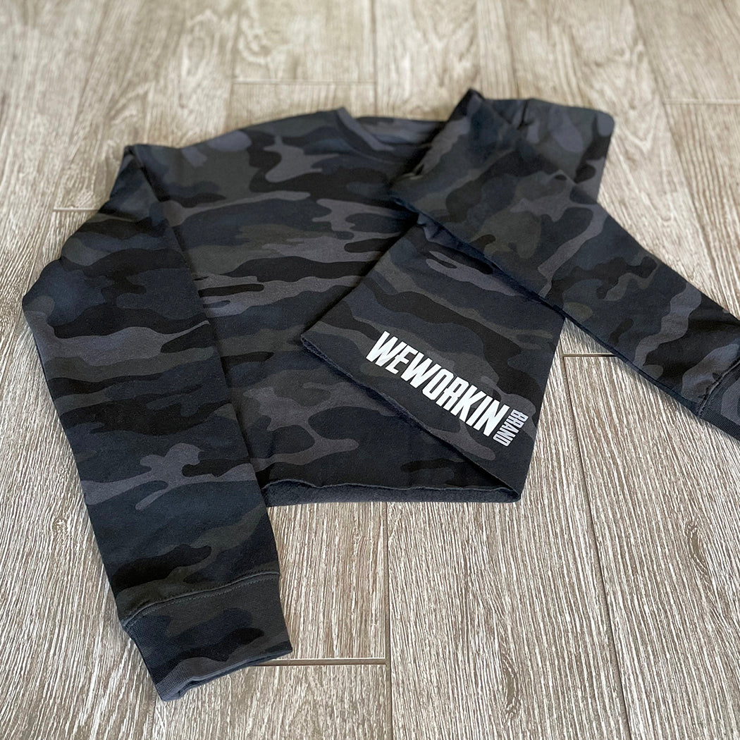 Front view of a We Workin women's lightweight cropped crew pullover in Black Camo, folded to show the back print (back print only, no front print). "WEWORKIN BRAND" block logo is printed on the back, bottom left hem area in white and grey ink. Imprint approx 5.5" wide.