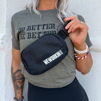 Woman wearing a WW Black crossbody belted bag pictured from front. WEWORKIN BRAND logo imprinted in white and grey ink on the front right lower edge. Smooth matte polyester material, zippered main compartment, color-matched metal zipper pulls on a molded zipper, color-matched interior pocket, with adjustable waist strap.